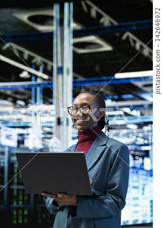 Cheerful female technician in data center using laptop to monitor rigs metrics. Jolly African american woman in server farm using notebook to enhance operational effectiveness of infrastructure 142646972