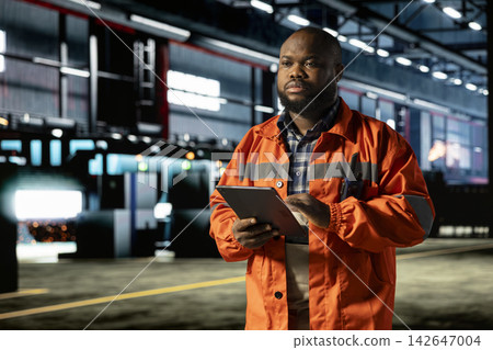 African american man manages heavy machinery activity on tablet in manufacturing facility, wearing protective gear and walking in the factory. Labor strength for engineering operations. African american man manages heavy machinery activity on tablet in manufacturing facility, wearing protective gear and walking in the factory. Labor strength for engineering operations. 142647004