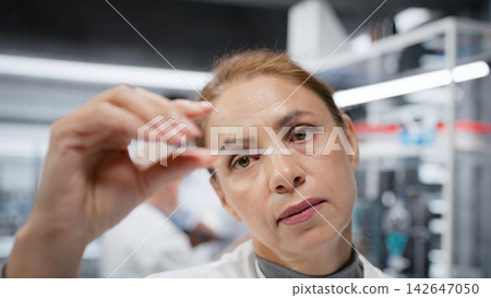 Materials scientist inspecting thin glass slide for structural defects or impurities. Research employee conducting visual quality control check using compound microscope, camera B 142647050