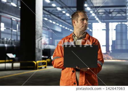 Worker performing inspection over fabrication tasks on laptop, wearing safety gear inside heavy industry warehouse. Engineering skills and the power of industrial machinery for production. Worker performing inspection over fabrication tasks on laptop, wearing safety gear inside heavy industry warehouse. Engineering skills and the power of industrial machinery for production. 142647063