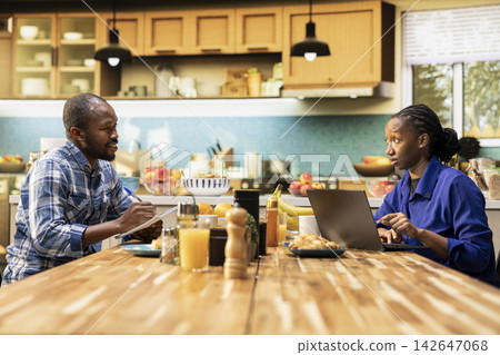 Young black couple planning grocery orders online using a laptop. Boyfriend and girlfriend discuss shopping list and comparing supermarket prices, choosing items for consumption needs. 142647068