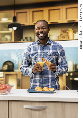 African American man serving fresh orange juice and a croissant in the kitchen, making breakfast. Smiling joyous guy enjoying organic homemade food, healthy morning routine and lifestyle. African American man serving fresh orange juice and a croissant in the kitchen, making breakfast. Smiling joyous guy enjoying organic homemade food, healthy morning routine and lifestyle. 142647075