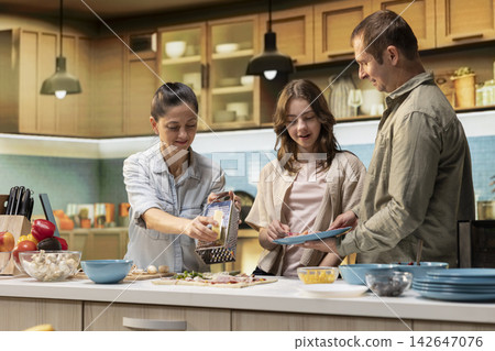 Pre-teen daughter sprinkling grated cheese in the kitchen and helping parents prepare pizza, family cooking activity during the weekend. Cozy moment of teamwork and meal preparation. 142647076
