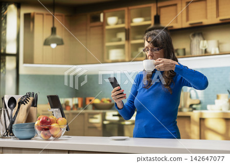 Mature person standing in home kitchen enjoying joyful moment of retirement. Pensioner with candid smile and relaxed posture reflecting natural wellbeing and positive household lifestyle. 142647077