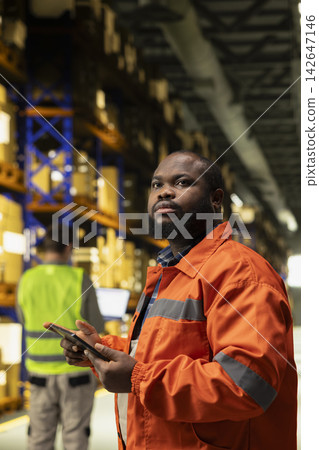 African american male overseeing cargo details on tablet in busy logistics hub, safety vest and professional workflow. Employee handling packages and merchandise for global e-commerce. African american male overseeing cargo details on tablet in busy logistics hub, safety vest and professional workflow. Employee handling packages and merchandise for global e-commerce. 142647146