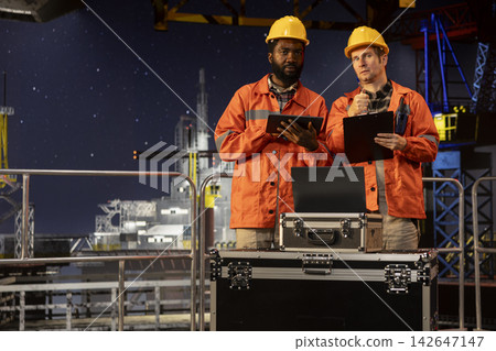 Diverse engineers colleagues using walkie talkie communication on marine oil platform, assessing fuel extraction data to maintain balance of natural gas and petroleum from the ocean habitat. Diverse engineers colleagues using walkie talkie communication on marine oil platform, assessing fuel extraction data to maintain balance of natural gas and petroleum from the ocean habitat. 142647147