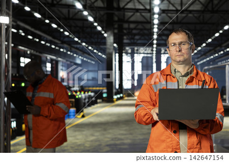 Professional employee in warehouse plant using laptop to guide operator teams through assembly tasks, demonstrating engineering and consistent manufacturing process activity. Automation. Professional employee in warehouse plant using laptop to guide operator teams through assembly tasks, demonstrating engineering and consistent manufacturing process activity. Automation. 142647154