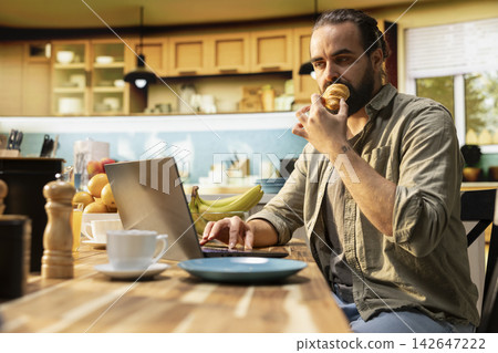Remote worker, eating croissants, using laptop in kitchen, solving daily tasks online, serving breakfast. Digital nomad, freelancer lifestyle with flexible work, multitasking and teleworking. Remote worker, eating croissants, using laptop in kitchen, solving daily tasks online, serving breakfast. Digital nomad, freelancer lifestyle with flexible work, multitasking and teleworking. 142647222