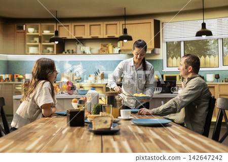 Pre-teen daughter sitting together with parents at breakfast table, serving scrambled eggs with bacon and pastry. Cozy scene captures bonding time during weekend morning routine. Pre-teen daughter sitting together with parents at breakfast table, serving scrambled eggs with bacon and pastry. Cozy scene captures bonding time during weekend morning routine. 142647242