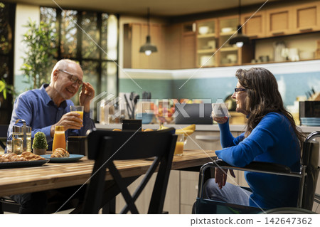 Mature woman with chronic disability serving coffee and pastry with her partner, enjoying cozy morning routine with laughter and casual conversation. Positive household moment in modern home. Mature woman with chronic disability serving coffee and pastry with her partner, enjoying cozy morning routine with laughter and casual conversation. Positive household moment in modern home. 142647362