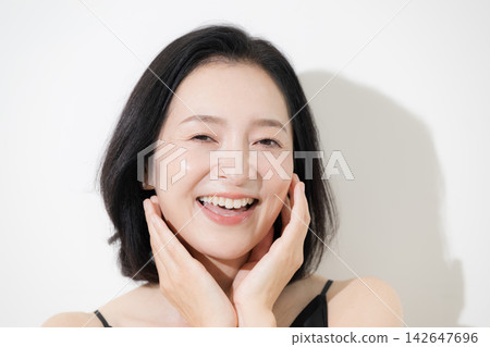 一位50歲左右、皮膚完美無瑕的美麗女性對著鏡頭燦爛地笑著，一邊撫摸著臉頰，一邊談論著護膚和抗衰老。 142647696