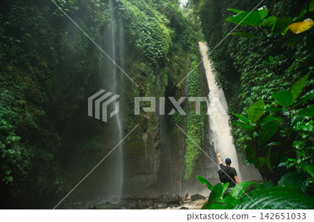 A traveler admires a tall waterfall in lush greenery A traveler admires a tall waterfall in lush greenery 142651033