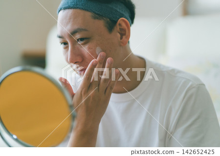A 40-year-old man applying lotion to his cheeks while looking in the mirror: skincare routine A 40-year-old man applying lotion to his cheeks while looking in the mirror: skincare routine 142652425