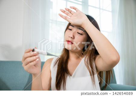 Sick Asian young woman sit under blanket on sofa she sneeze with digital thermometer 142654089