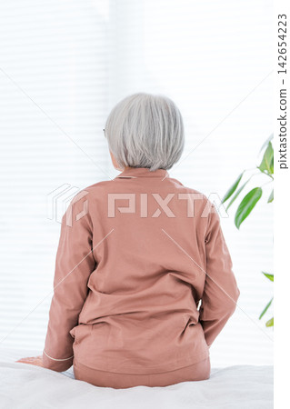 一位老婦人在清晨醒來,坐在床上,背影清晰可見。畫面展現了她晚年獨立健康的生活。 一位老婦人在清晨醒來,坐在床上,背影清晰可見。畫面展現了她晚年獨立健康的生活。 142654223