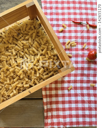Top view of raw ditalini pasta in a wooden crate with cherry tomatoes and chili peppers on a red checkered tablecloth Top view of raw ditalini pasta in a wooden crate with cherry tomatoes and chili peppers on a red checkered tablecloth 142655170