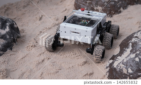 Compact wheeled rover moves across soft sand beside dark rocks, showing rugged engineering, off-road mobility, and experimental robotics for research, testing, exploration, and innovation projects Compact wheeled rover moves across soft sand beside dark rocks, showing rugged engineering, off-road mobility, and experimental robotics for research, testing, exploration, and innovation projects 142655832