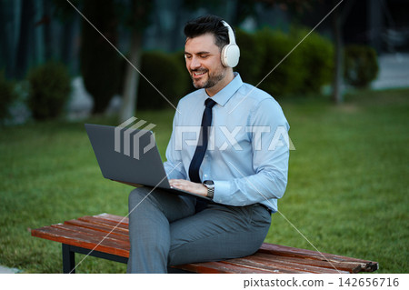 Mid adult man smiling while telecommuting from a park bench Mid adult man smiling while telecommuting from a park bench 142656716