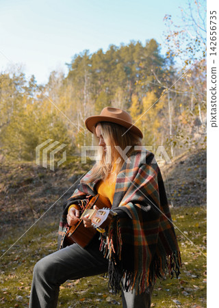 Beautiful blonde girl playing acoustic guitar and singing relaxing enjoying outdoors on sunny autumn day. Hippie woman in boho style learning and practicing playing musical instrument. Fall leaves Beautiful blonde girl playing acoustic guitar and singing relaxing enjoying outdoors on sunny autumn day. Hippie woman in boho style learning and practicing playing musical instrument. Fall leaves 142656735