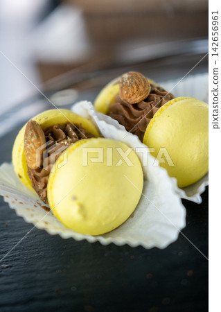 Almond meringue. Buttercream piping. Yellow macarons in paper package display on dark tray. Sweet pastry with choco filling served in cafeteria. Almond meringue. Buttercream piping. Yellow macarons in paper package display on dark tray. Sweet pastry with choco filling served in cafeteria. 142656961