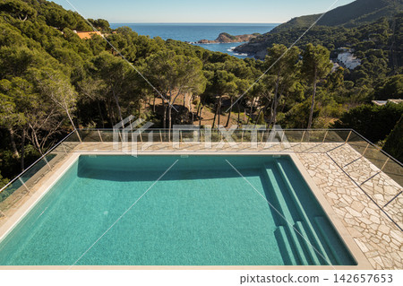 Clear rectangular pool with shallow steps overlooking tree lined hillside and distant ocean under bright sunlight beside stone deck, lush greenery, scenic resort setting Clear rectangular pool with shallow steps overlooking tree lined hillside and distant ocean under bright sunlight beside stone deck, lush greenery, scenic resort setting 142657653
