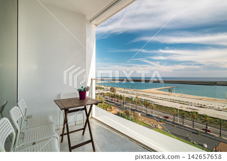 Modern balcony with white chairs and wooden table overlooking beach, ocean, blue sky, clouds, glass railing, potted plant, relaxing coastal resort setting, with balcony, chair, table, wooden detail Modern balcony with white chairs and wooden table overlooking beach, ocean, blue sky, clouds, glass railing, potted plant, relaxing coastal resort setting, with balcony, chair, table, wooden detail 142657658