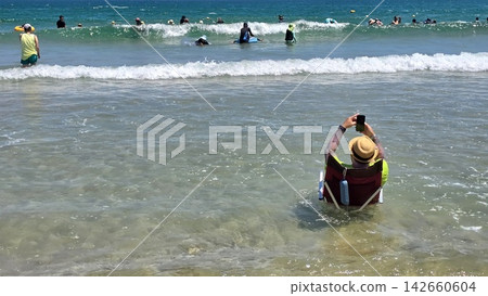 "A man taking a selfie in a swimming ring on a bustling beach," "A summer beach crowded with swimmers," "A man holding his smartphone at the water's edge." "A man taking a selfie in a swimming ring on a bustling beach," "A summer beach crowded with swimmers," "A man holding his smartphone at the water's edge." 142660604