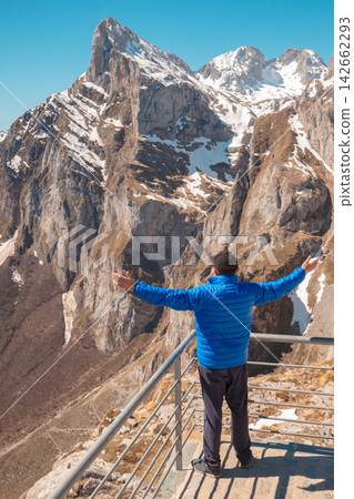 A man with hands in the air on a viewpoint against mountains covered with snow. A man looks at the beautiful panorama of the mountains. National Park Peaks of Europe. Fuente De, Cantabria, Spain A man with hands in the air on a viewpoint against mountains covered with snow. A man looks at the beautiful panorama of the mountains. National Park Peaks of Europe. Fuente De, Cantabria, Spain 142662293