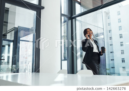 Happy woman, office and business discussion with phone call by window for proposal or friendly chat. Young female person or financial advisor talking on mobile smartphone with coffee for conversation Happy woman, office and business discussion with phone call by window for proposal or friendly chat. Young female person or financial advisor talking on mobile smartphone with coffee for conversation 142663276