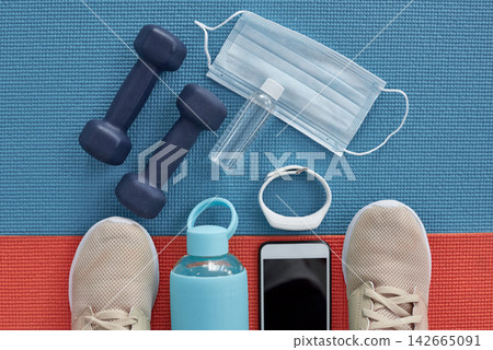 Workout, equipment and face mask on yoga mat with tech for fitness tracker and gym progress. Above, floor and wellbeing for illness prevention at health and wellness club for sport with weights Workout, equipment and face mask on yoga mat with tech for fitness tracker and gym progress. Above, floor and wellbeing for illness prevention at health and wellness club for sport with weights 142665091