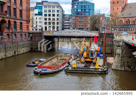 Yellow hydraulic excavator on a construction barge performing restoration works inside the canal of the historic Speicherstadt district in Hamburg, Germany 142665995