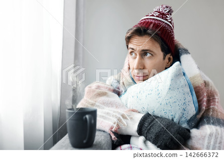 cold man deals with a home heating problem during a harsh cold snap and energy crisis, wrapped in a blanket with a hot drink to cope with weak heating and frost. cold man deals with a home heating problem during a harsh cold snap and energy crisis, wrapped in a blanket with a hot drink to cope with weak heating and frost. 142666372