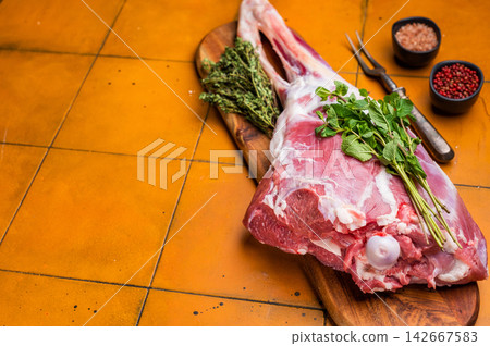 Whole raw lamb leg, uncooked mutton meat on butcher board. orange background. top view Whole raw lamb leg, uncooked mutton meat on butcher board. orange background. top view 142667583