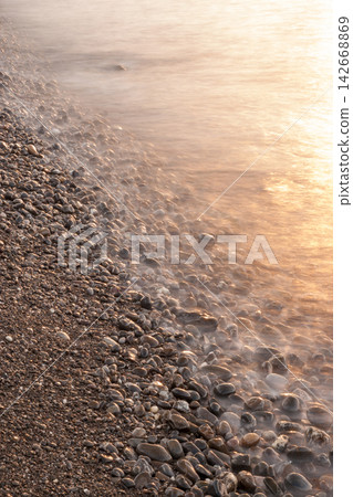 Long exposure vertical shot of gentle sunset wave flowing over pebble beach Long exposure vertical shot of gentle sunset wave flowing over pebble beach 142668869