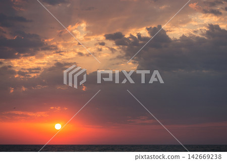 Dramatic sunset over sea horizon with glowing red sun and dark clouds 142669238