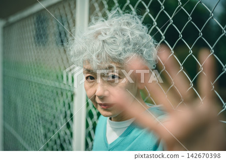 一位老婦人透過鐵絲網凝視著我們,臉上帶著憂鬱的神情。 一位老婦人透過鐵絲網凝視著我們,臉上帶著憂鬱的神情。 142670398