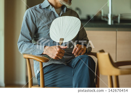 一位老人在家裡的椅子上放鬆休息,用風扇為自己降溫。 一位老人在家裡的椅子上放鬆休息,用風扇為自己降溫。 142670454