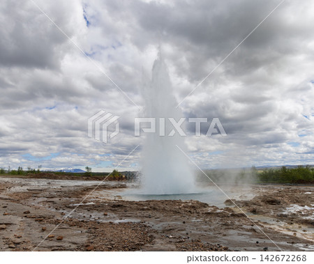 Strokkur geyser erupts from a blue pool in Haukadalur under layered clouds Strokkur geyser erupts from a blue pool in Haukadalur under layered clouds 142672268
