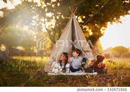 Teepee, kids and fun in garden tent outdoor in nature for camping, playing and adventure with pointing. Cloth, light and fantasy house for children in forest with imagination, suitcase and teddy bear Teepee, kids and fun in garden tent outdoor in nature for camping, playing and adventure with pointing. Cloth, light and fantasy house for children in forest with imagination, suitcase and teddy bear 142672888