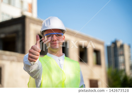 Construction professional in hard hat giving thumbs up at an active building site. Project approval client satisfaction and successful construction progress Construction professional in hard hat giving thumbs up at an active building site. Project approval client satisfaction and successful construction progress 142673528