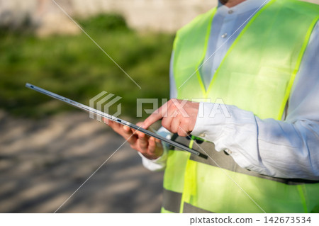 Construction professional using a tablet while wearing high visibility vest at an outdoor worksite. Digital construction management field inspection and paperless site documentation Construction professional using a tablet while wearing high visibility vest at an outdoor worksite. Digital construction management field inspection and paperless site documentation 142673534