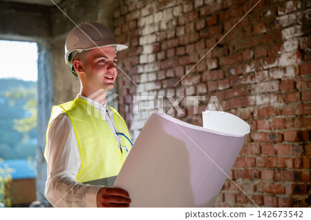 Construction professional reviewing architectural blueprints inside an unfinished brick building. Building design renovation planning and site coordination Construction professional reviewing architectural blueprints inside an unfinished brick building. Building design renovation planning and site coordination 142673542
