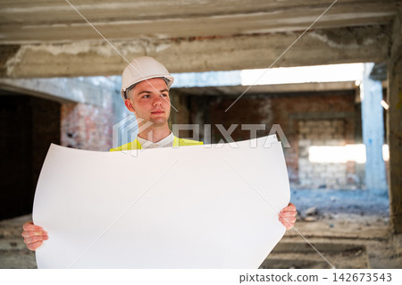 Construction professional holding large architectural plans inside an unfinished renovation building. Construction planning blueprint review and architectural design verification Construction professional holding large architectural plans inside an unfinished renovation building. Construction planning blueprint review and architectural design verification 142673543
