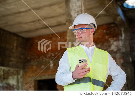 Architect using smartphone while wearing safety gear at an unfinished construction site. Mobile field reporting site coordination and digital construction workflow Architect using smartphone while wearing safety gear at an unfinished construction site. Mobile field reporting site coordination and digital construction workflow 142673545