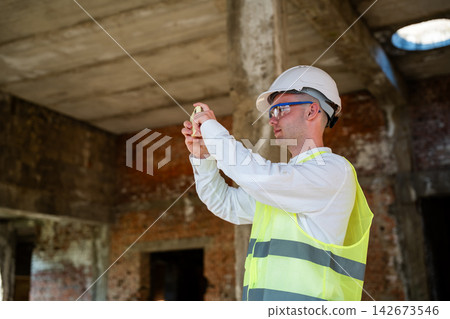 Construction professional using smartphone inside unfinished building site while wearing safety equipment. Digital construction management remote inspection and progress reporting Construction professional using smartphone inside unfinished building site while wearing safety equipment. Digital construction management remote inspection and progress reporting 142673546