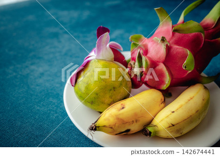 A white plate adorned with tropical fruits: dragon fruit, banana, lime, and orchids, against a blue fabric background. A white plate adorned with tropical fruits: dragon fruit, banana, lime, and orchids, against a blue fabric background. 142674441