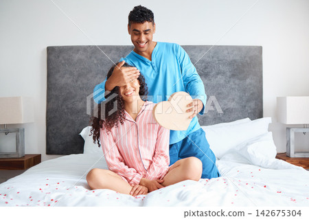 Surprise, valentines day and couple on bed for happiness, chocolate gift or romance in bedroom. Heart shape, smile and married people in home for anniversary, present or bonding together in morning Surprise, valentines day and couple on bed for happiness, chocolate gift or romance in bedroom. Heart shape, smile and married people in home for anniversary, present or bonding together in morning 142675304
