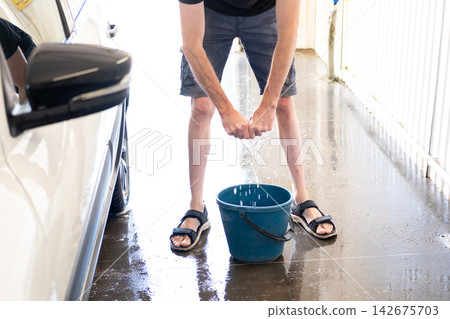 Man wringing out cloth into blue plastic bucket at car wash. High angle view of a man in shorts and sandals wringing out a wet rag into a blue bucket on a wet concrete floor next to a white car Man wringing out cloth into blue plastic bucket at car wash. High angle view of a man in shorts and sandals wringing out a wet rag into a blue bucket on a wet concrete floor next to a white car 142675703