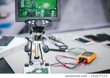 Digital microscope inspecting a green circuit board on an electronics repair workbench with probes, tools, smartphone parts, and a yellow multimeter. Digital microscope inspecting a green circuit board on an electronics repair workbench with probes, tools, smartphone parts, and a yellow multimeter. 142677123