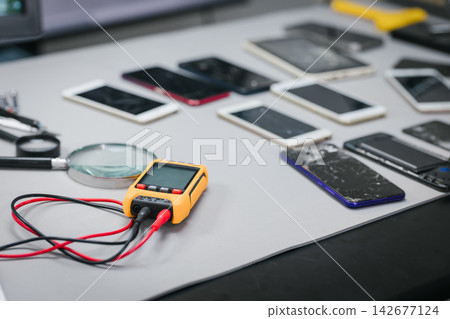 Smartphones with cracked screens on an electronics repair bench beside a digital multimeter, test leads, and magnifying glass. Smartphones with cracked screens on an electronics repair bench beside a digital multimeter, test leads, and magnifying glass. 142677124